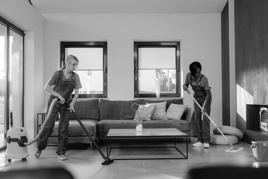 Two professional cleaners from Harringay Cleaner are performing surface cleaning in a modern living room. They are wearing matching grey shirts and red overalls, each holding a vacuum or mop. The room