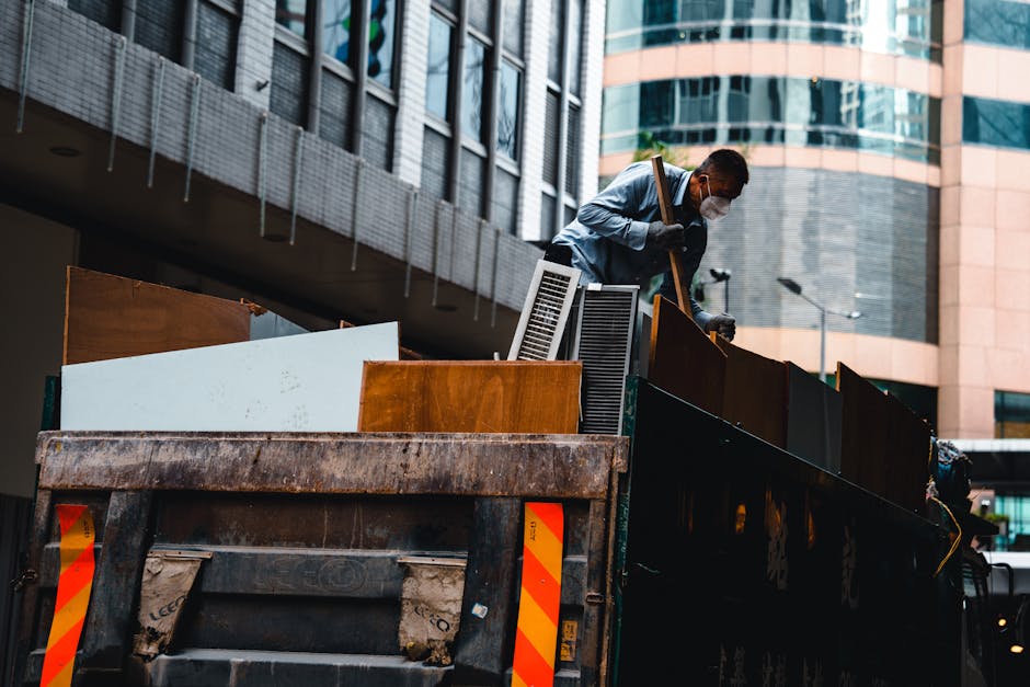 A worker wearing a face mask and gloves is standing on the edge of a large black truck loaded with various discarded items, including wooden panels, metal fixtures, and cabinets, during daytime. The w