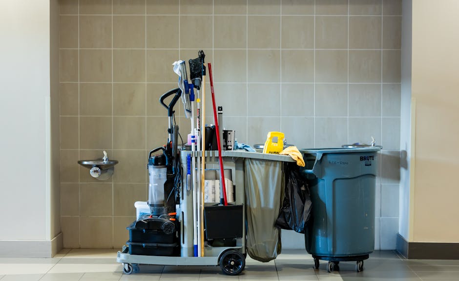 A worker wearing a face mask and gloves is standing on the edge of a large black truck loaded with various discarded items, including wooden panels, metal fixtures, and cabinets, during daytime. The w