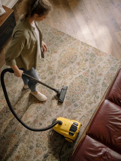 A woman vacuuming a beige, patterned carpet in a living room. The carpet features intricate floral designs in shades of green, orange, and beige, and is situated beside a red leather sofa. She is dressed casually in a light brown jacket, white sneakers, and jeans, and is using a yellow vacuum cleaner with a black hose. The room has wooden flooring bordering the carpet and is well-lit, emphasizing the cleanliness and freshness of the surface. This scene illustrates domestic surface cleaning as part of Harringay Cleaner's professional carpet cleaning services near N4, Harringay Ladder, highlighting thorough and effective sanitisation.