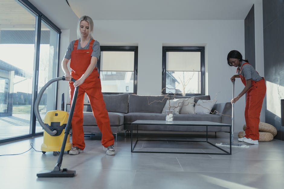 Two professional cleaners from Harringay Cleaner are performing surface cleaning and deep cleaning tasks in a modern living room. One cleaner is vacuuming the hardwood floor with a canister vacuum cle