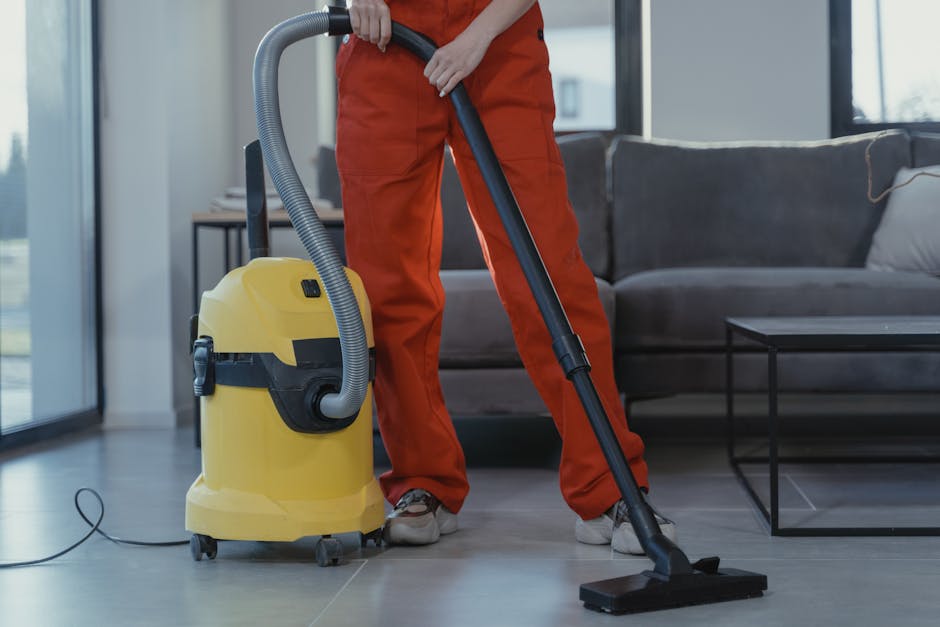A person using a yellow vacuum cleaner with a black hose on a patterned area rug in a living room. The individual is wearing a light-colored jacket, fitted jeans, and white sneakers, and is leaning fo